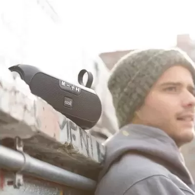 Coluna de som portátil preta sobre parede de tijolos com jovem de gorro cinzento ao fundo