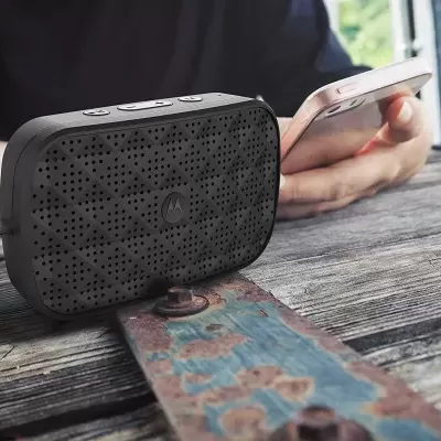 Coluna portátil preta com perfurações sobre mesa de madeira ao lado de smartphone