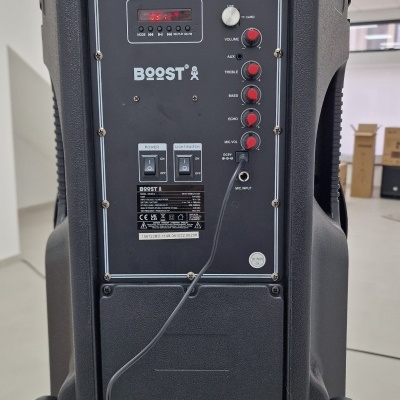 Coluna portátil preta BOOST com botões e rodas numa sala clara