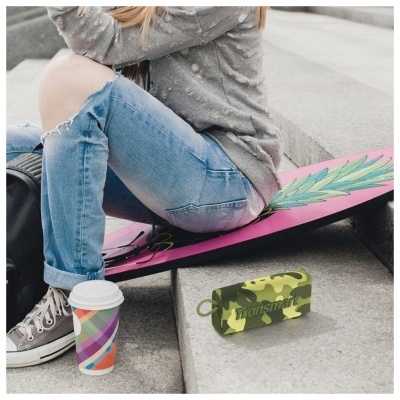 Pessoa sentada com calças de ganga rasgadas e ténis ao lado de caixa de som verde camuflada e copo de papel colorido