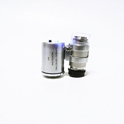 Microscópio portátil metálico prateado com lentes e texto 'CURRENCY DETECTING MICROSCOPE 60X' sobre fundo branco