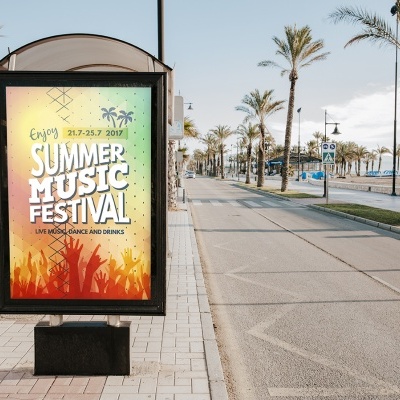 Cartaz de festival de música de verão num expositor urbano alinhado em avenida com palmeiras e praia ao lado.
