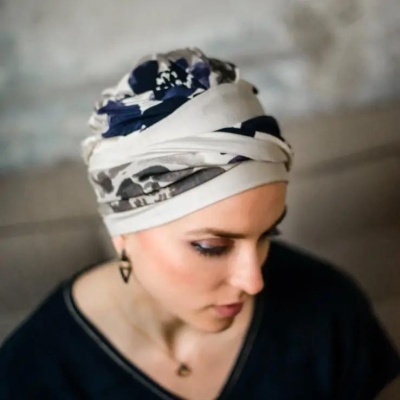 Pessoa com lenço de cabeça estampado em tons de branco, cinza, azul escuro, camisola preta e brincos triangulares.