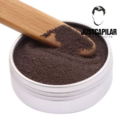 Pote branco com pó escuro e espátula de madeira ao lado de texto JUSTCAPILAR SYSTEM