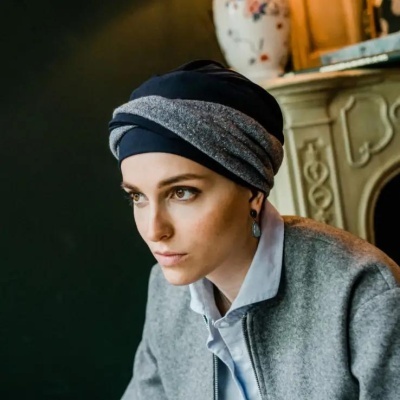 Mulher com turbante cinzento e preto e casaco cinzento perto de lareira