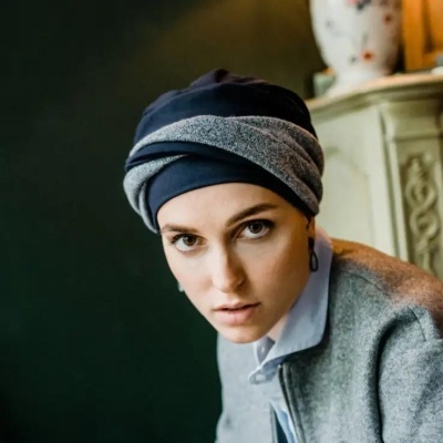 Turbante azul escuro com faixa cinzenta usado por mulher com camisa azul clara e casaco cinzento num ambiente interior.