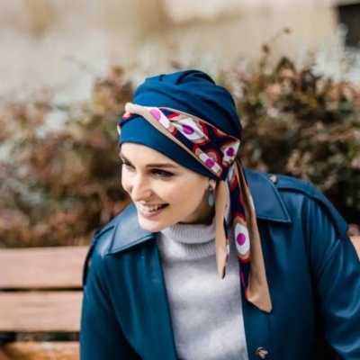 Mulher com lenço azul escuro estampado na cabeça, casaco azul e camisola cinza sentada num banco ao ar livre