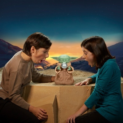 Duas crianças olham surpreendidas para figura de Baby Yoda numa mesa quadrada bege.