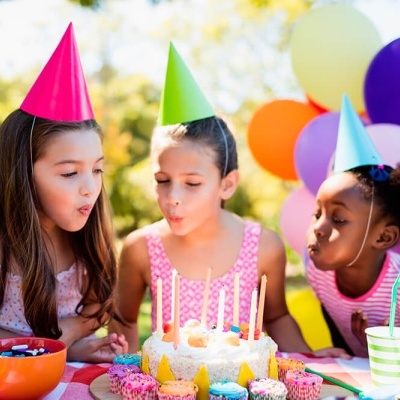 Três meninas com chapéus de festa a soprar velas num bolo branco ao ar livre