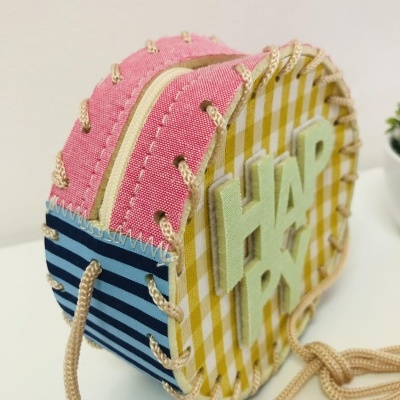 Bolsa redonda colorida com padrão listrado e xadrez, com a palavra HAPPY em relevo