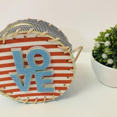 Cesta redonda com padrão claro e letras azuis LOVE, ao lado de planta artificial num vaso branco.