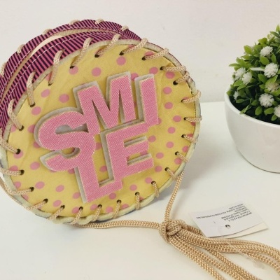 Mala redonda amarela com padrão de bolinhas cor-de-rosa e texto 'SMILE' em rosa, ao lado vaso branco com planta artificial