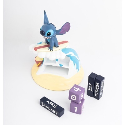 Figura brinquedo azul tipo Stitch sobre prancha de surf com base de areia e ondas e blocos de calendário