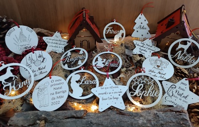 Decorações de Natal em madeira branca com nomes e texto em português, decoradas com detalhes vermelhos, sobre fundo de madeira com luzes.