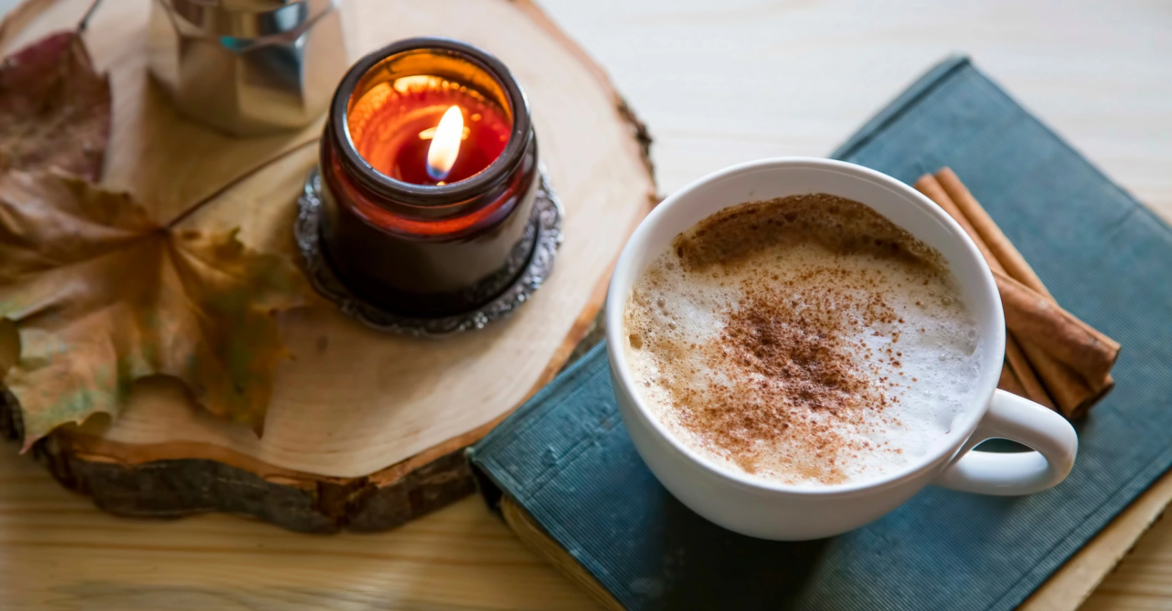 Chávena branca com bebida cremosa ao lado de vela acesa em frasco castanho sobre base de madeira e folha seca