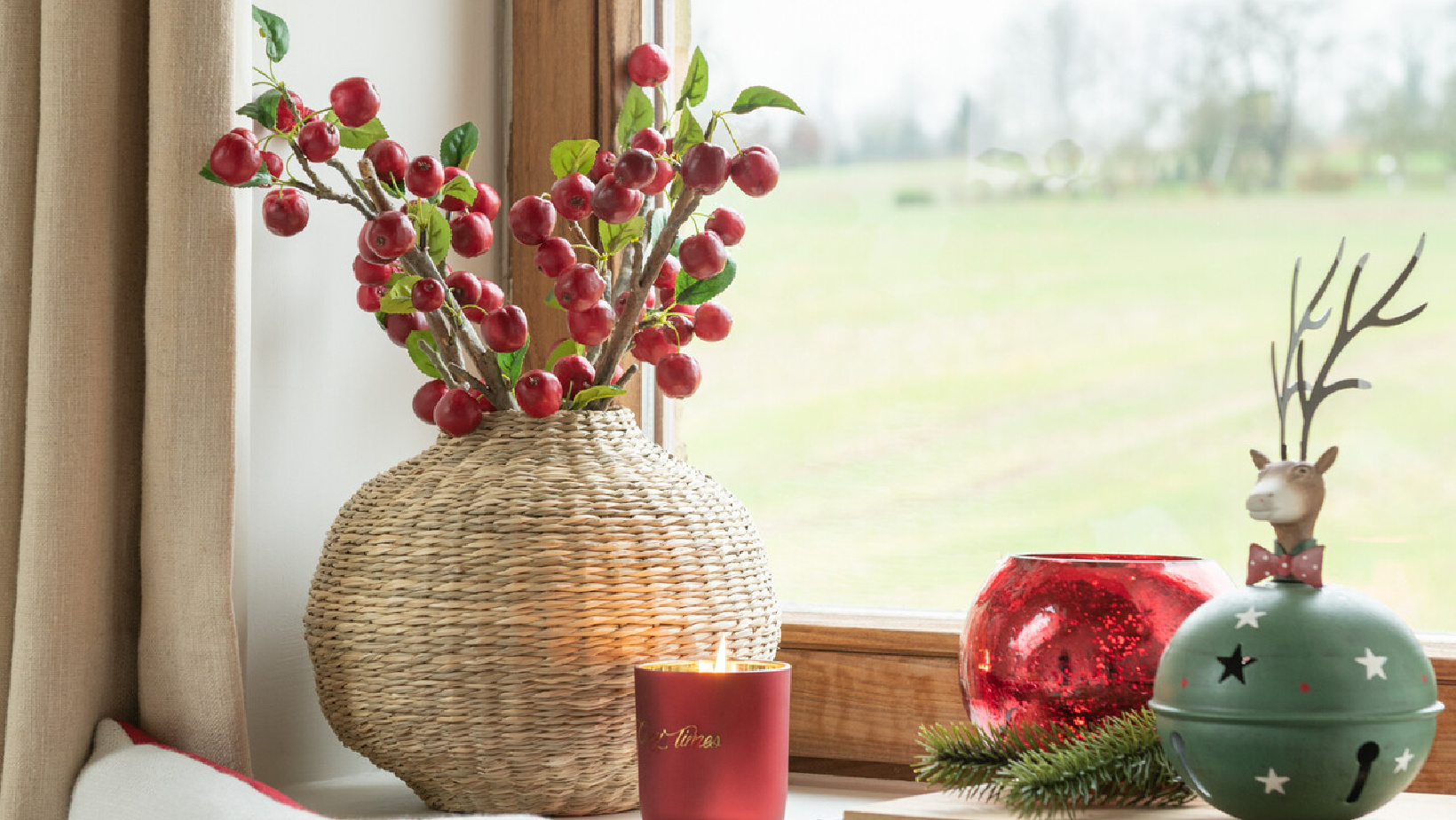 Vaso decorativo de fibra natural com ramos de frutas vermelhas, vela vermelha, bola vermelha e bola verde com cervo numa janela