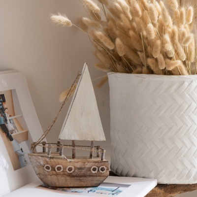 Barco decorativo em madeira com vela branca ao lado de vaso branco com flores secas