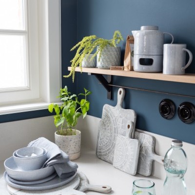 Utensílios de cozinha em cerâmica cinza, tábuas de madeira brancas com padrão, plantas verdes, jarra e copo de vidro sobre bancada branca com parede azul