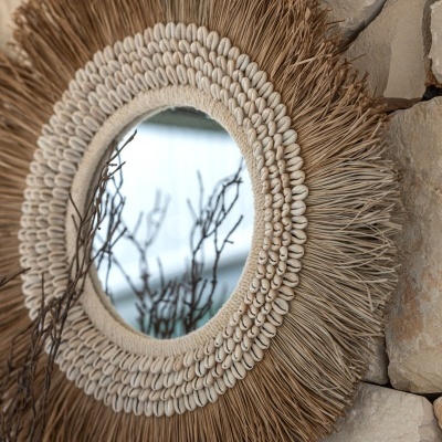 Espelho redondo com moldura de fibras naturais e conchas pendurado em parede de pedra.