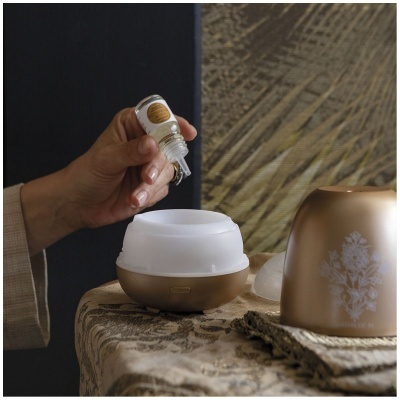 Difusor aromático dourado e branco numa mesa com mão a despejar óleo essencial de um pequeno frasco.