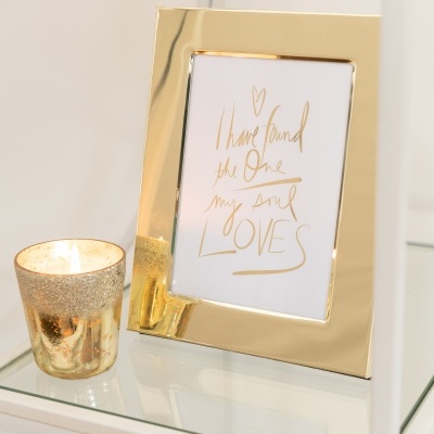 Moldura dourada com frase manuscrita e castiçal dourado com vela numa prateleira de vidro