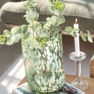 Vaso de vidro verde com flores secas ao lado de castiçal transparente com vela acesa sobre mesa de madeira