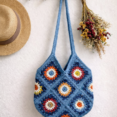 Bolsa de crochet azul com padrão colorido pendurada em suporte dourado na parede ao lado de chapéu e flores secas