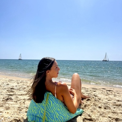 Mala de praia azul e verde em areia junto ao mar com mulher sentada ao lado