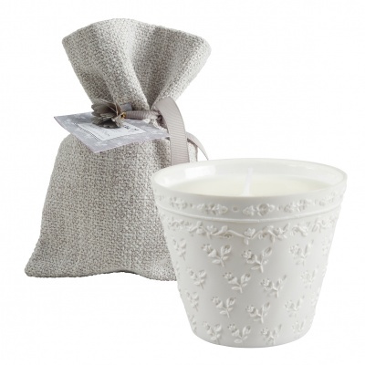 Vela branca em recipiente cerâmico com relevo floral ao lado de saco tecido cinza claro