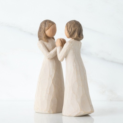 Duas figuras femininas esculpidas em madeira, vestidos creme, mãos dadas.