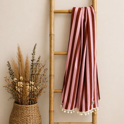 Manta riscada vermelha e rosa com pompons pendurada em escada de bambu, vaso com plantas secas ao lado
