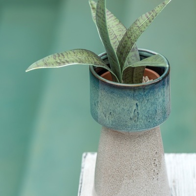 Planta artificial em vaso azul e pedestal cinzento sobre superfície branca