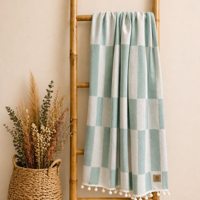 Manta verde água e branca com padrão quadriculado pendurada em suporte de madeira ao lado de cesta com plantas secas.