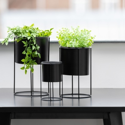 Três vasos de metal preto com plantas verdes artificiais em suportes finos de metal preto sobre mesa preta