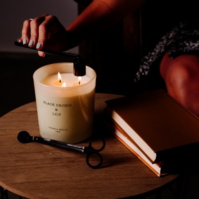 Vela perfumada com três pavios acesos num recipiente de vidro branco sobre mesa de madeira com livros e acendedor preto