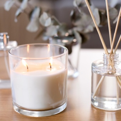 Vela branca acesa, difusor de varetas e frasco pequeno numa mesa de madeira clara