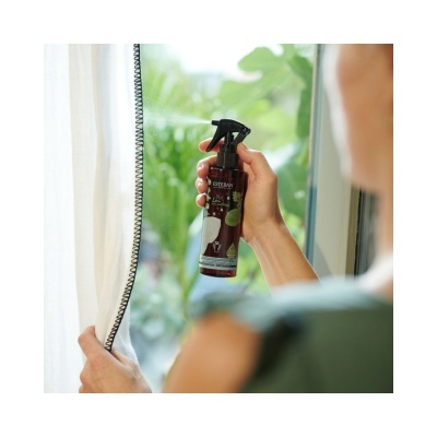 Frasco de spray marrom com tampa preta na mão junto a cortina branca e janela com plantas