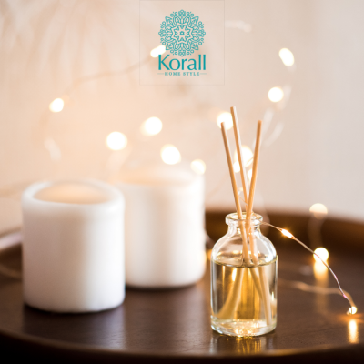 Frasco com varetas difusoras e velas brancas numa superfície de madeira escura com luzes decorativas e logotipo Korall no fundo.