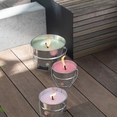 Três velas coloridas em recipientes metálicos com alças no chão de madeira junto a banco de madeira e blocos de cimento