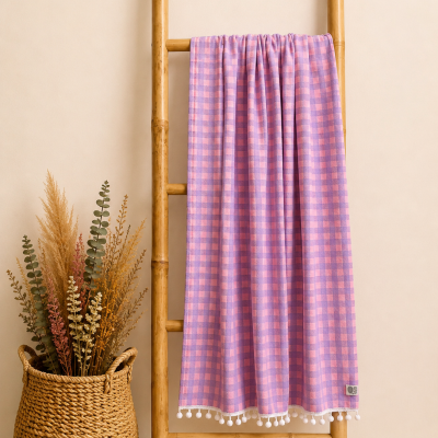 Manta xadrez rosa e lilás com pompons brancos pendurada em escada de bamboo ao lado de cesto de ráfia com flores secas