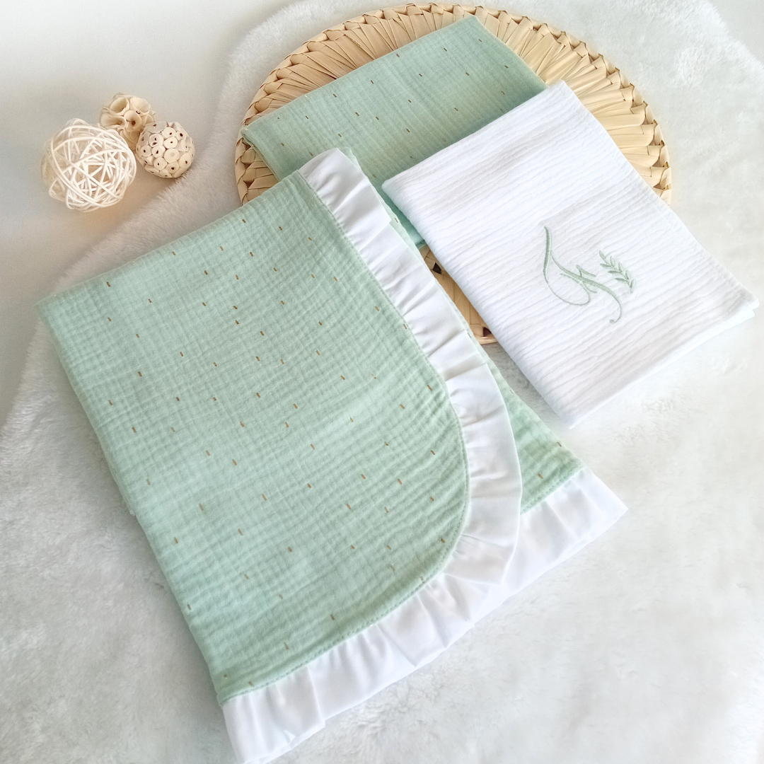 Conjunto de manta e fralda para bebé em algodão com cores verde água e branco, decorados e dispostos sobre base trançada.