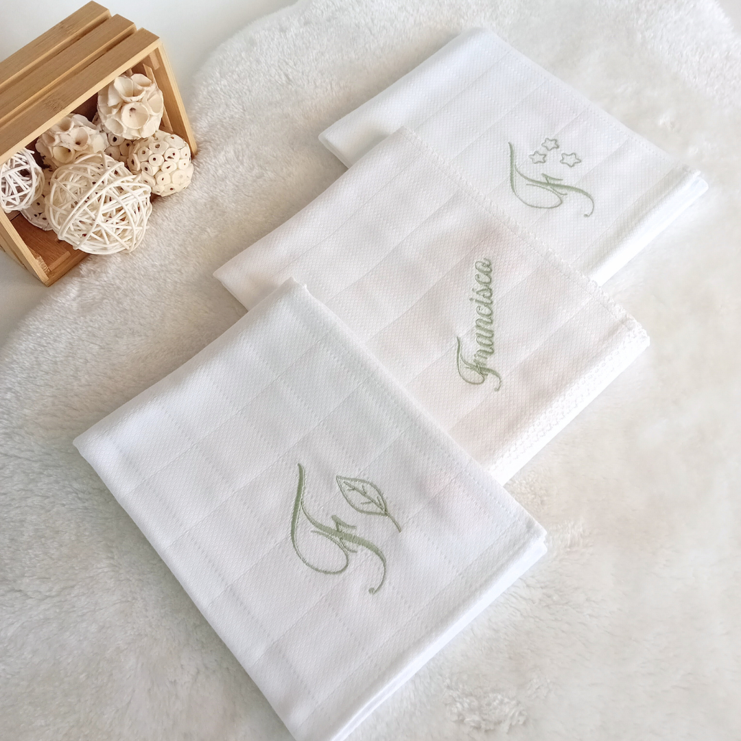 Três toalhas brancas bordadas dobradas sobre tapete branco com caixa de madeira e esferas decorativas