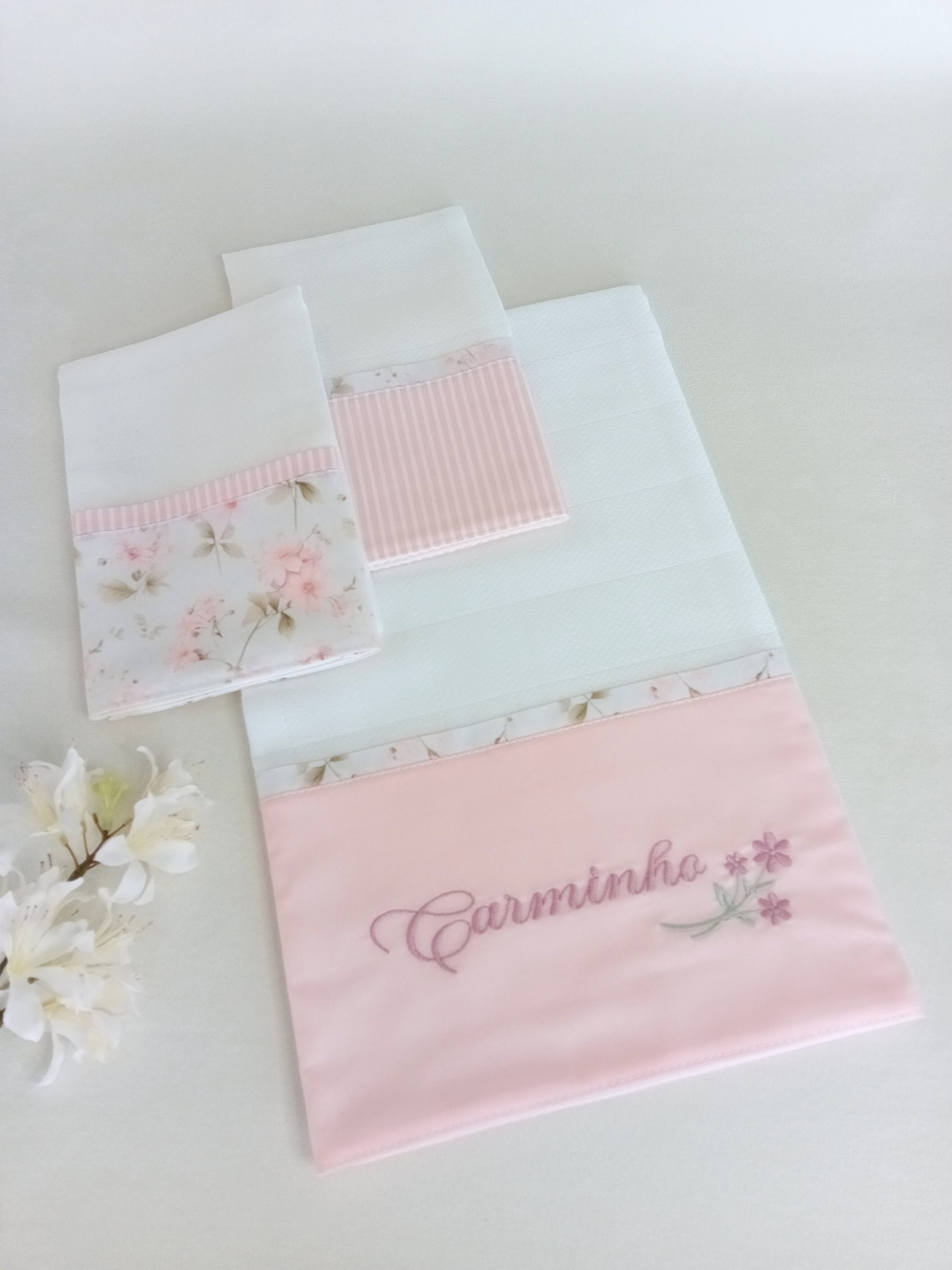 Fraldas de tecido branco com detalhes florais e bordado rosa com o texto Carminho.
