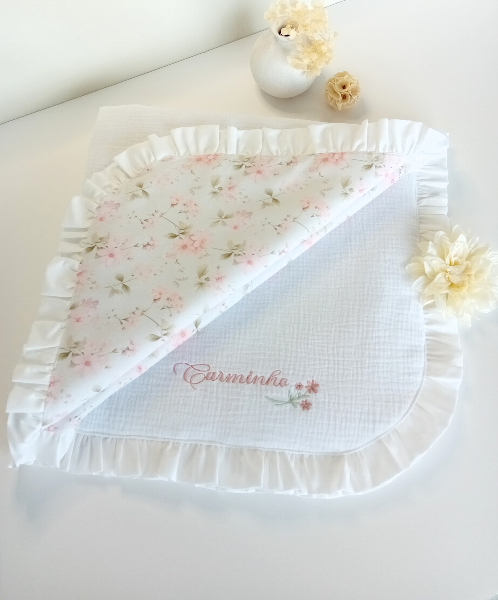 Manta branca com borda de folhos e padrão floral, com o texto bordado Carminho.