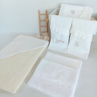 conjunto de roupa de bebé branca com bordados bege e toalha felpuda ao lado, mini escada de madeira ao fundo