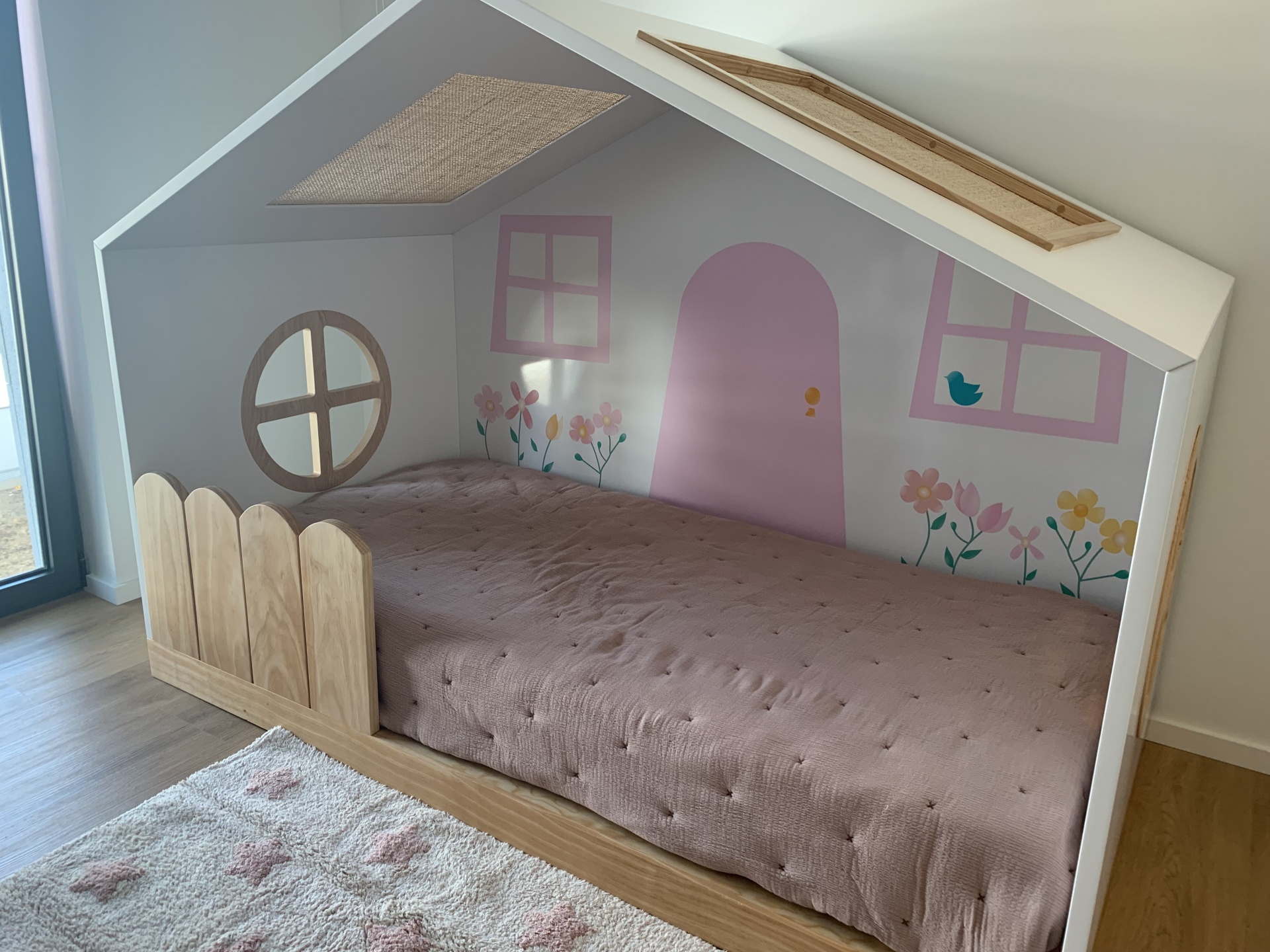 Cama infantil em forma de casinha com decoração floral e roupa de cama rosa clara.