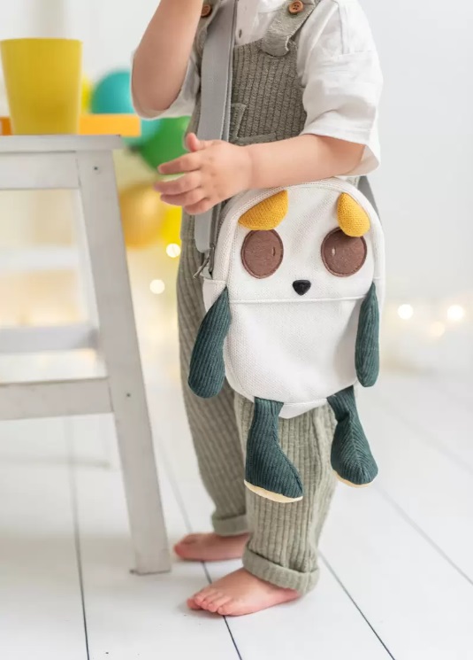 Mala Cuki Mochila infantil em forma de animal com detalhes amarelos, castanhos e verdes, usada por criança ao lado de mesa branca