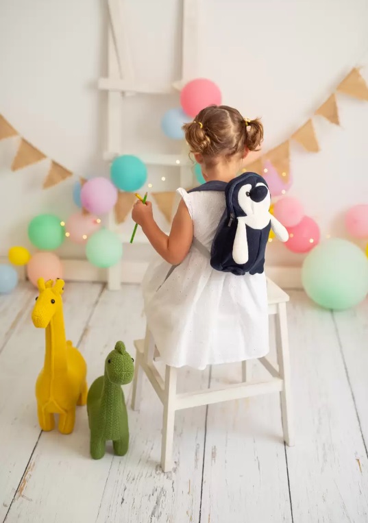 Mochila Pingui Criança com mochila de pinguim sentada em cadeira rodeada de balões e brinquedos em ambiente branco