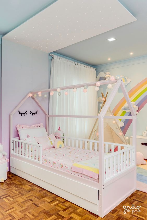 CAMA CABANINHA Cama infantil em madeira branca com decoração de pompons de luz e roupa de cama em tons pastéis num quarto colorido.
