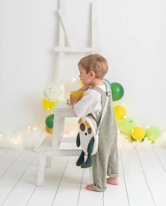 Mala Cuki Criança com jardineira e brinquedo de pelúcia em ambiente branco com balões.